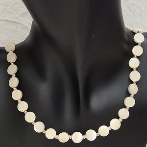 May include: A cream-colored necklace featuring round, flat beads. The beads are connected by small, silver-colored links. The necklace is displayed on a black mannequin, showcasing its design and shape. The necklace is a jewelry item.