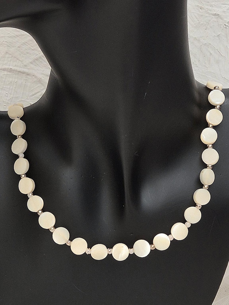 May include: A cream-colored necklace featuring round, flat beads. The beads are connected by small, silver-colored links. The necklace is displayed on a black mannequin, showcasing its design and shape. The necklace is a jewelry item.