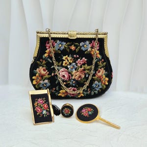 Puede incluir: Un bolso negro con cadena y marco dorados, con un diseño floral de punto de cruz. Los accesorios a juego incluyen un bolígrafo, un estuche, gemelos y un espejo de mano, todos con detalles florales y acentos dorados.