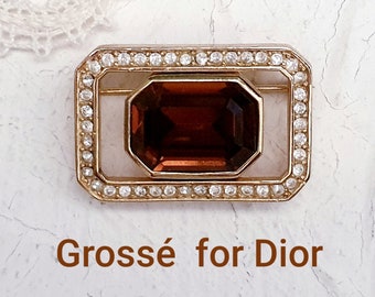 May include: A gold-toned brooch with a large, dark brown, faceted gemstone set in a square frame. The frame is surrounded by a row of clear rhinestones. The brooch is labeled "Grossé for Dior".