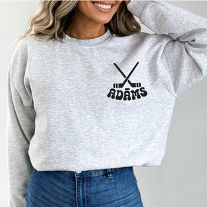 May include: A light grey sweatshirt with a crew neck. The sweatshirt displays a black graphic of crossed hockey sticks and the name "ADAMS" in a retro font. The design is centered on the chest.