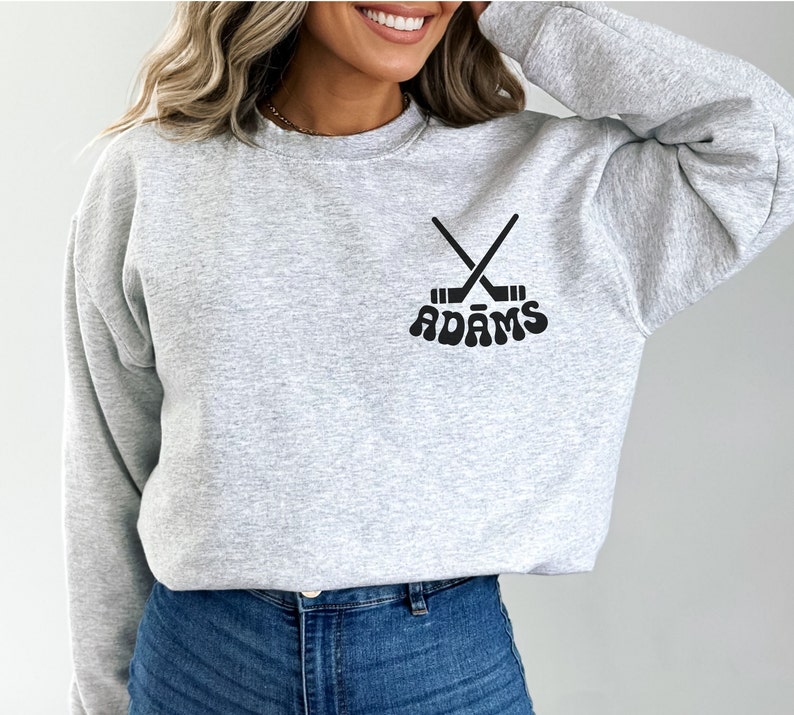 May include: A light grey sweatshirt with a crew neck. The sweatshirt displays a black graphic of crossed hockey sticks and the name "ADAMS" in a retro font. The design is centered on the chest.