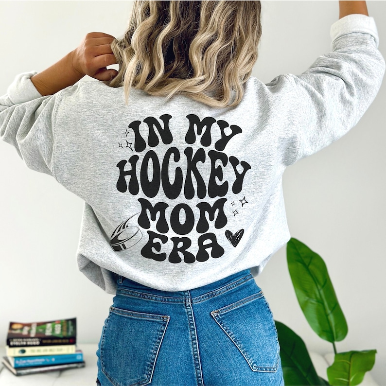 May include: Light grey sweatshirt featuring the black text "IN MY HOCKEY MOM ERA" in a retro font, with a hockey puck graphic. The sweatshirt is worn with blue jeans.