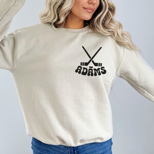 May include: A beige crew neck sweatshirt featuring a black hockey stick design and the name "ADAMS" in a retro font. The sweatshirt is a neutral colour, suitable for casual wear. The design is centered on the chest.