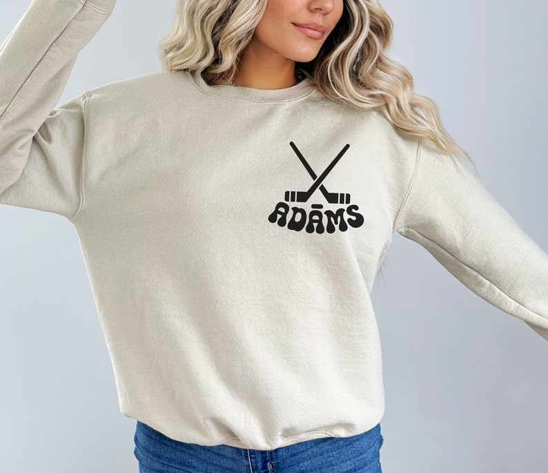 May include: A beige crew neck sweatshirt featuring a black hockey stick design and the name "ADAMS" in a retro font. The sweatshirt is a neutral colour, suitable for casual wear. The design is centered on the chest.