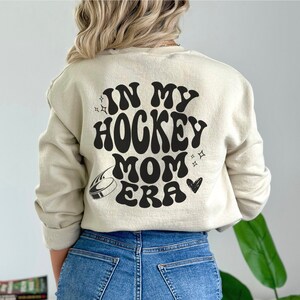 May include: A cream-coloured sweatshirt featuring the phrase "IN MY HOCKEY MOM ERA" in black, retro font. The design includes a hockey puck, stars, and a heart. The sweatshirt has a crew neck and is a neutral colour.