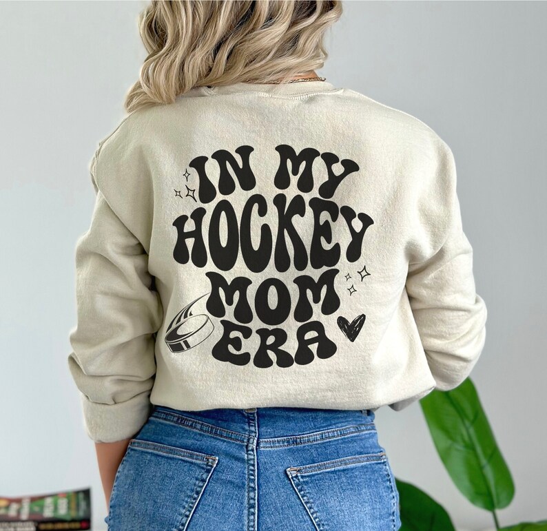 May include: A cream-coloured sweatshirt featuring the phrase "IN MY HOCKEY MOM ERA" in black, retro font. The design includes a hockey puck, stars, and a heart. The sweatshirt has a crew neck and is a neutral colour.