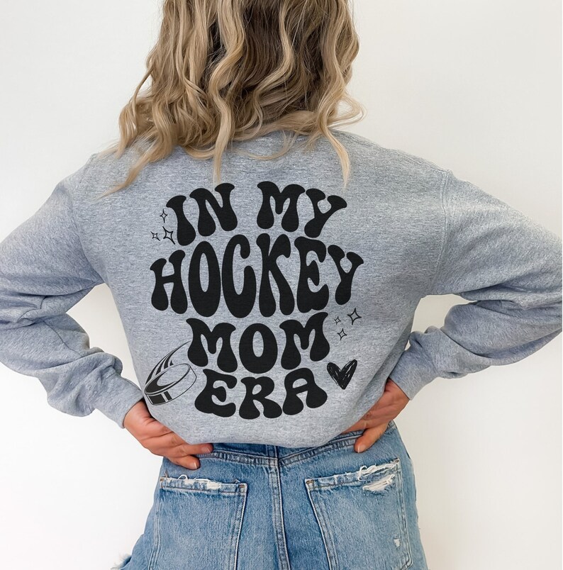 May include: A grey sweatshirt featuring the black text "IN MY HOCKEY MOM ERA" in a retro font. The design includes a hockey puck, stars, and a heart. The sweatshirt is worn with blue denim jeans.