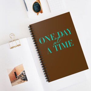 Puede incluir: Una libreta espiral marrón con el texto "ONE DAY at a TIME" en letras azul turquesa.