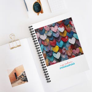 Puede incluir: Un cuaderno de espiral con una cubierta colorida que presenta un patrón de corazones pintados en varios tonos de rojo, azul, amarillo, verde y morado. El texto "my sober journal" está impreso en la cubierta en rosa y azul.