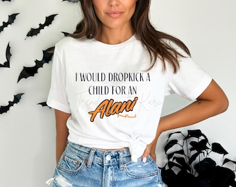 May include: White t-shirt with the text "I WOULD DROPKICK A CHILD FOR AN Alani Nu". The Alani Nu logo is in orange and black. Black bat decorations are on the wall in the background.