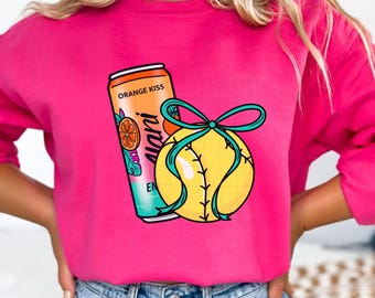 May include: A bright pink sweatshirt featuring a graphic of a yellow softball with teal stitching and a teal bow, next to an orange energy drink can with the text "Orange Kiss" and "Alani Nu".