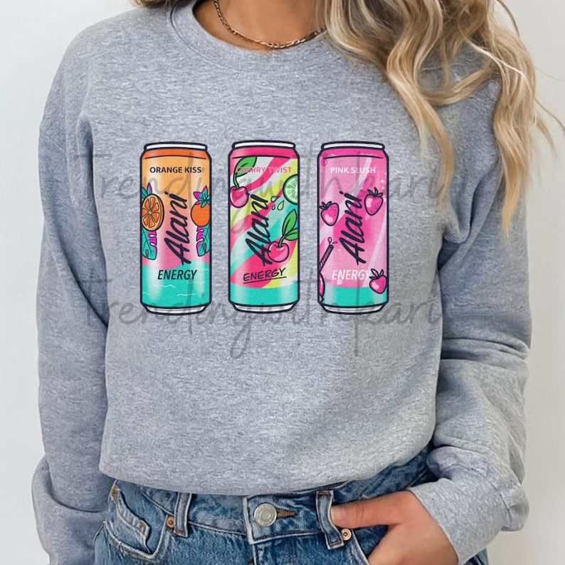 May include: A heather gray sweatshirt featuring three colorful energy drink cans. The cans are labeled "Orange Kiss," "Berry Twist," and "Pink Slush," with the brand name "Alani" prominently displayed. Each can has a unique design with fruit illustrations.