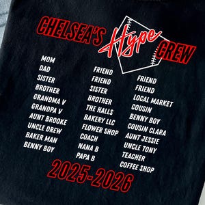 Può includere: T-shirt nera con la scritta "CHELSEA'S Hype CREW" in rosso e bianco. La maglietta elenca nomi come Mamma, Papà e Amico, insieme a "2025-2026" in rosso. È presente anche una grafica di baseball.