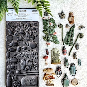 May include: A silicone mould for crafting with a variety of nature-themed designs, including mushrooms, beetles, ferns, and a glass cloche. The mould is grey and has the text "Decor Moulds Midnight Muses" printed on it.