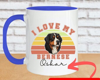 May include: White ceramic coffee mug with a blue rim and handle. The mug has a retro sunset design with the text "I love my Bernese Oskar" and a cartoon image of a Bernese Mountain Dog.