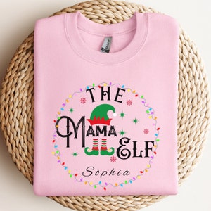 May include: A light pink sweatshirt with the text "The Mama Elf" in a circular design. The design includes an elf hat, snowflakes, and colorful lights. The name "Sophia" is printed below the text.