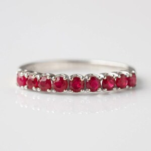 May include: A silver ring set with a line of bright red gemstones. The gemstones are round and closely set, forming a continuous band of colour. The ring is shown against a white backdrop, emphasising its elegant design.