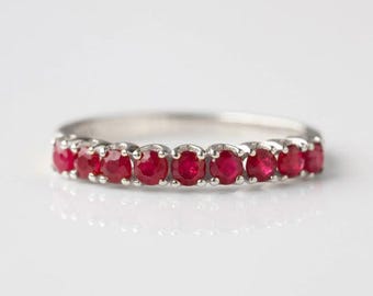 May include: A silver ring featuring a row of vibrant red gemstones. The gemstones are round-cut and set closely together, creating a continuous band of color. The ring is displayed on a white background, highlighting its elegant design.