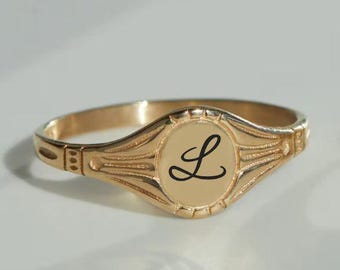 May include: A gold-colored signet ring with an oval face. The face features the cursive letter "L" in black. The ring has a decorative band with etched details. The ring is set against a light background.