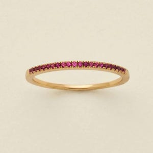 May include: A delicate gold ring with a band of small, vibrant pink gemstones. The ring has a thin, rounded band and the gemstones are set closely together, creating a continuous line of color. The ring is displayed against a neutral background.