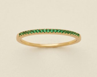 Puede incluir: Un anillo de banda dorado con una fila de pequeñas piedras preciosas de color verde brillante. El anillo se presenta sobre un fondo neutro, resaltando el contraste entre el oro y las piedras verdes. Una joya delicada.