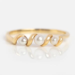 May include: A gold ring with a unique design featuring three white pearls. The ring's band has a swirling pattern, with each pearl nestled within the curves of the gold. The ring is set against a white background.