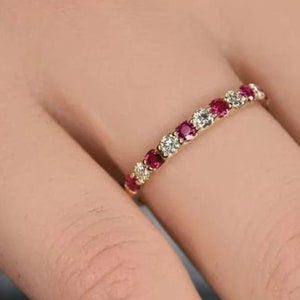 May include: A gold ring featuring alternating round-cut red and clear gemstones. The ring is displayed on a finger, highlighting the sparkle of the stones. The design of the ring is a classic and elegant style.