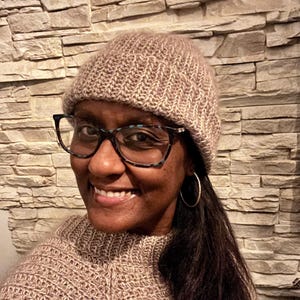 May include: A knitted beanie and sweater in a light brown color. The beanie is worn on the head, and the sweater is worn on the upper body. The person is wearing glasses and hoop earrings. The background is a stone wall.