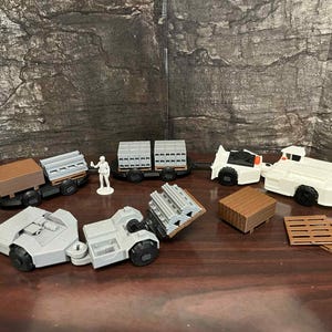 May include: A collection of toy construction vehicles and accessories, including trucks, trailers, and pallets. The vehicles are primarily white and gray, with brown accents. A small figurine is also present.