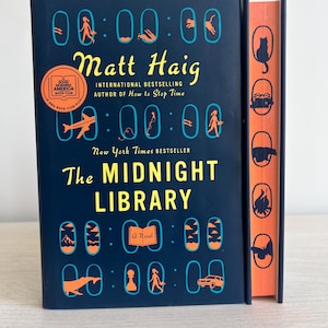 May include: A dark blue hardcover book with the title "The Midnight Library" by Matt Haig. The book cover features a repeating pattern of clocks with various images inside each clock.
