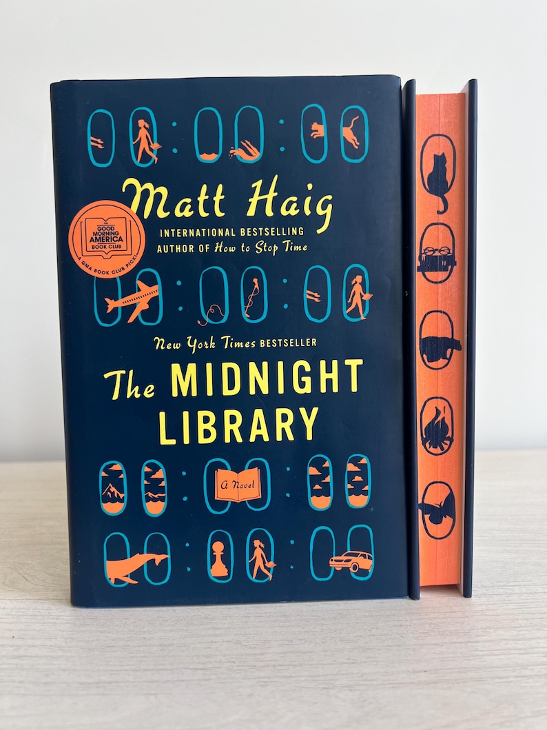 May include: A dark blue hardcover book with the title "The Midnight Library" by Matt Haig. The book cover features a repeating pattern of clocks with various images inside each clock.