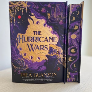 Op de afbeelding: Een hardback boek in paars en goud met de titel "The Hurricane Wars: A Novel" van Thea Guanzon. De omslag toont een kaart met een schip en een halve maan.