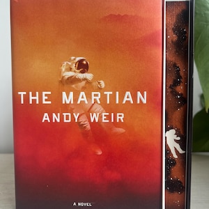 May include: A book cover with a red and orange background featuring an astronaut floating in space. The title "The Martian" and the author "Andy Weir" are printed in white on the cover. The book is a novel.