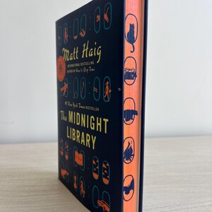 May include: A dark blue hardcover book with a black and white illustration of a clock on the cover. The title "The Midnight Library" is in white text. The author's name "Matt Haig" is in yellow text. The book's spine is orange with black silhouettes of various objects.