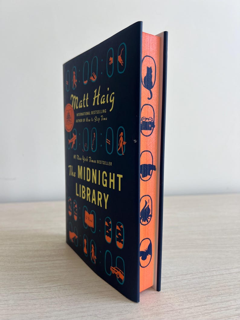 May include: A dark blue hardcover book with a black and white illustration of a clock on the cover. The title "The Midnight Library" is in white text. The author's name "Matt Haig" is in yellow text. The book's spine is orange with black silhouettes of various objects.