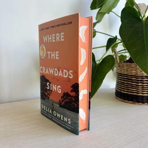 May include: A paperback book titled "Where the Crawdads Sing" by Delia Owens. The cover features a sunset scene with a silhouette of a person walking on a beach. The book is a New York Times bestseller.