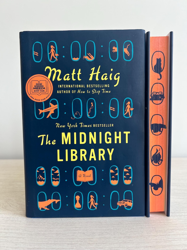 May include: A blue hardcover book with the title "The Midnight Library" by Matt Haig. The book is a New York Times bestseller and a Good Morning America Book Club pick. The cover features a series of clocks with different symbols inside each clock.