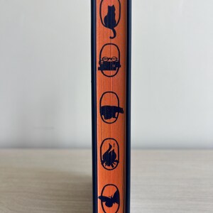 May include: The spine of a hardcover book with an orange cover and a dark blue border. The cover features five black silhouettes of a cat, a bowl of food, a pig, a spider, and a butterfly.