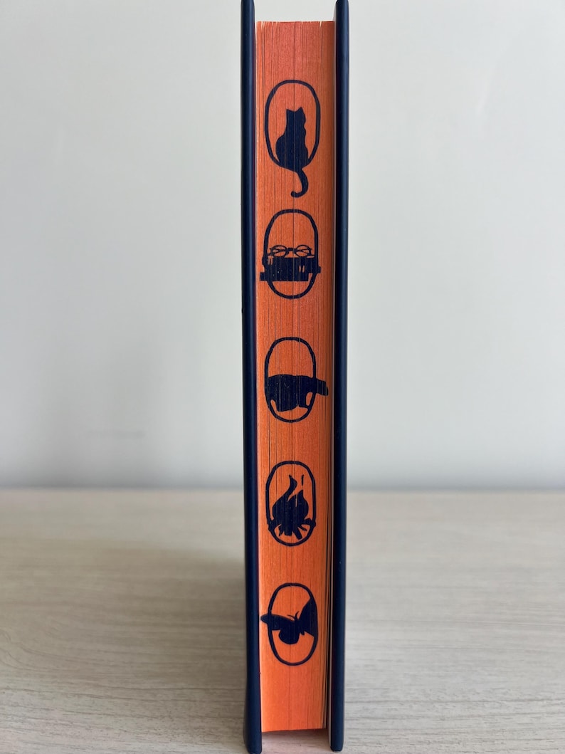 May include: The spine of a hardcover book with an orange cover and a dark blue border. The cover features five black silhouettes of a cat, a bowl of food, a pig, a spider, and a butterfly.