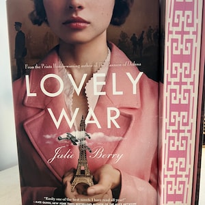 Puede incluir: Un libro titulado "LOVELY WAR" de Julie Berry. La portada presenta a una mujer con un abrigo rosa sosteniendo una Torre Eiffel en miniatura. Se ve el lomo de un segundo libro, con un patrón geométrico rosa y blanco.
