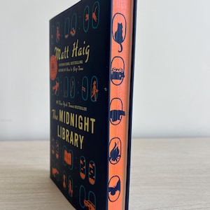 May include: A dark blue hardcover book with a black and white illustration of a cat on the spine. The title "The Midnight Library" is printed in yellow on the front cover. The author's name, "Matt Haig", is printed in yellow above the title.