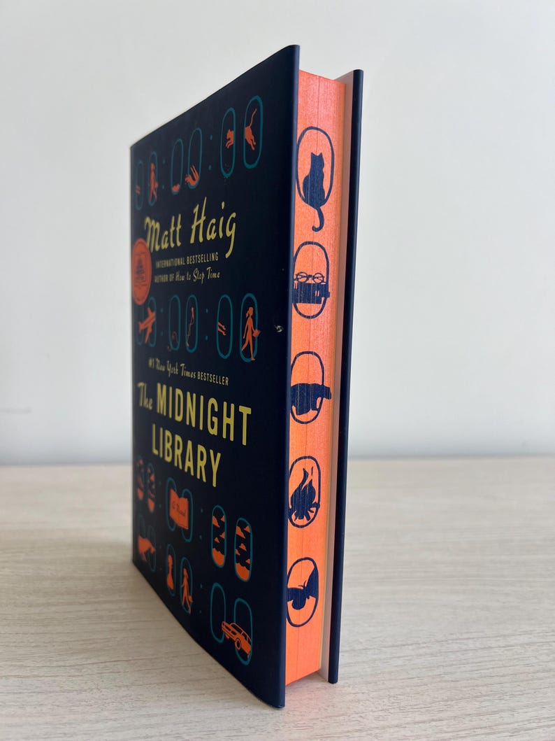 May include: A dark blue hardcover book with a black and white illustration of a cat on the spine. The title "The Midnight Library" is printed in yellow on the front cover. The author's name, "Matt Haig", is printed in yellow above the title.