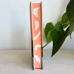 May include: The spine of a paperback book with a peach colored cover. The spine has a repeating pattern of white feathers.