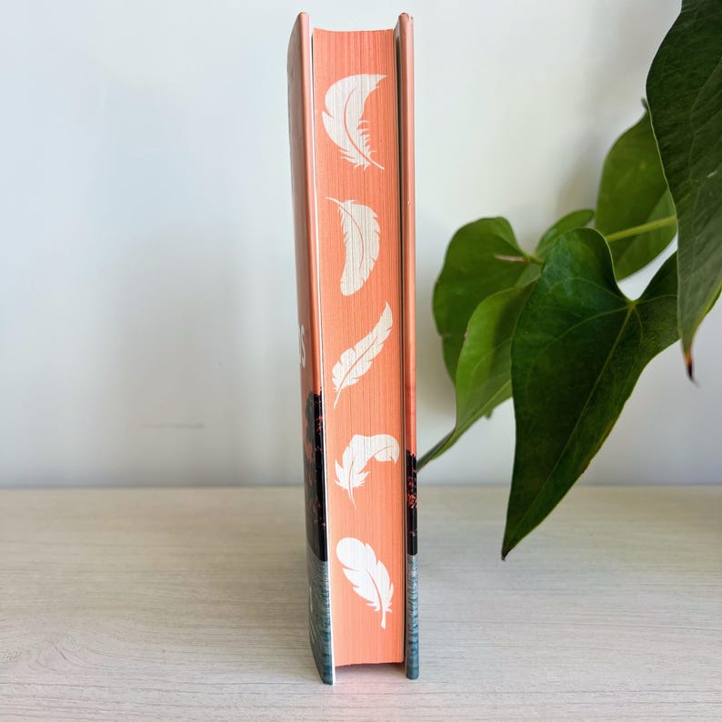 May include: The spine of a paperback book with a peach colored cover. The spine has a repeating pattern of white feathers.