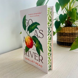 May include: A hardback book titled "Go As A River" by Shelley Read. The cover displays a peach branch with a ripe peach and green leaves. The spine has a green and white leaf pattern. The book is on a light-coloured surface.