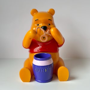 Peut inclure: Figurine jaune de Winnie l'ourson avec un t-shirt rouge, tenant un cure-miel. Un pot de miel bleu avec une bande rose et le mot "HUNNY" est devant la figurine. La figurine est assise.