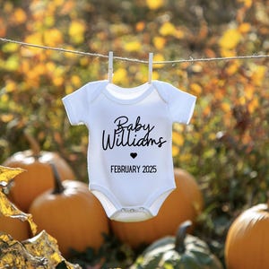 Pode incluir: Body de bebé branco com o texto "Baby Williams" e um símbolo de coração, juntamente com a data "Fevereiro 2025". O body está pendurado num varal com molas, num fundo de abóboras e folhagem de outono.