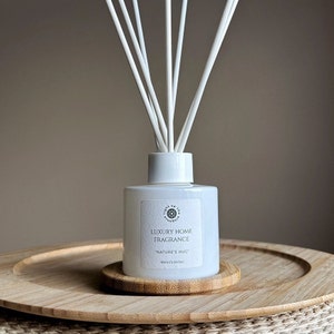 Könnte beinhalten: Weiße Diffuserflasche mit einem weißen Etikett, auf dem "Luxury Home Fragrance "Nature's Hug" 50ml/1.64oz" steht. Die Flasche steht auf einem Holzuntersetzer und hat weiße Diffuserstäbchen.