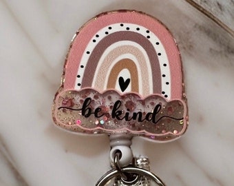May include: A pink and brown rainbow with a heart and glitter, with the words "be kind" written on it. This is a retractable badge holder.